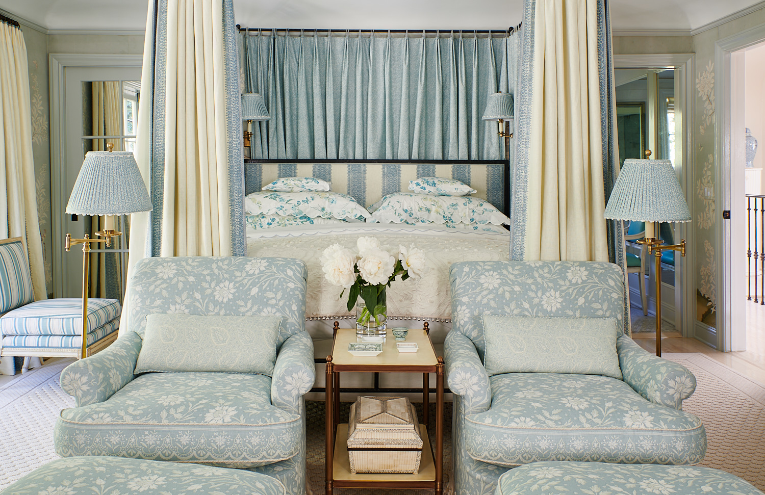 Wide view of bed drapes, stripe upholstered headboard, floral shams, and floral upholstered club chairs with matching ottomans